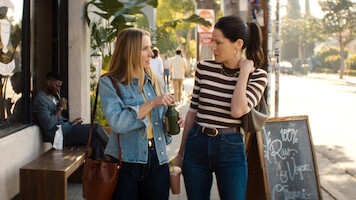 Two women talking on a sunny sidewalk outside a café with plants and a chalkboard sign. One woman holds a drink; both carry bags. A person sits on a bench nearby, looking at a phone. Urban street scene with passing pedestrians.