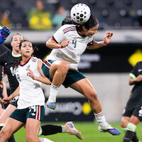 Female soccer player in white jersey jumps and heads the ball during a match; opponents and teammates are nearby on a professional field with blurred stadium background.