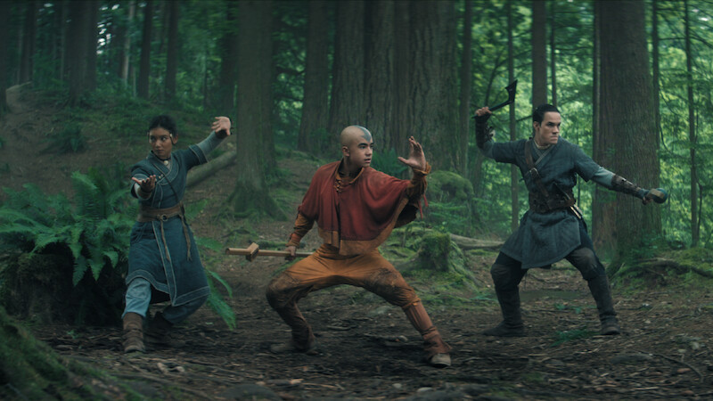Three people in martial arts stances, dressed in fantasy costumes, stand ready for action in a lush, green forest with tall trees and ferns, suggesting a dramatic, adventurous scene.