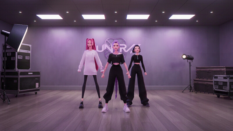Three animated women posing in a modern dance studio with wooden floors, gray walls, professional lighting, and gear cases visible, standing under bright lights.