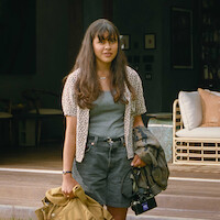 Young woman with long hair stands outside a cozy, modern house holding a coat, bag, and camera. The relaxed setting features a wicker sofa and bookshelves, evoking a calm, casual, and welcoming atmosphere.