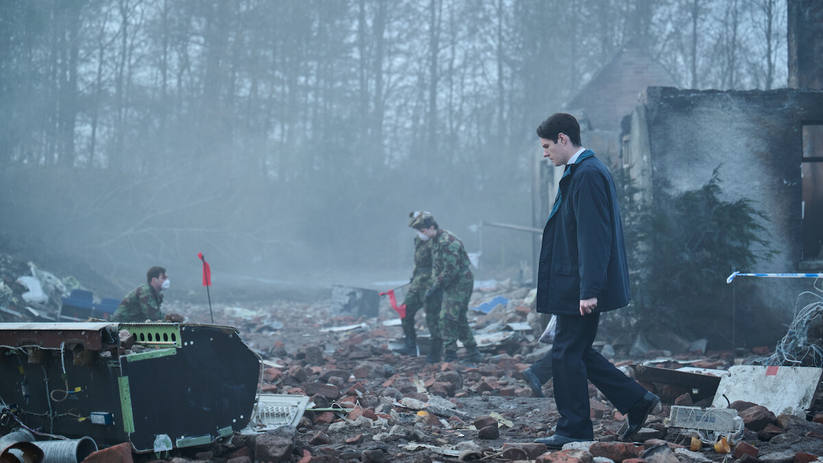 A man in a suit walks through a debris-strewn, foggy ruin as two people in camouflage uniforms investigate the wreckage; trees and a partially destroyed building are in the background.