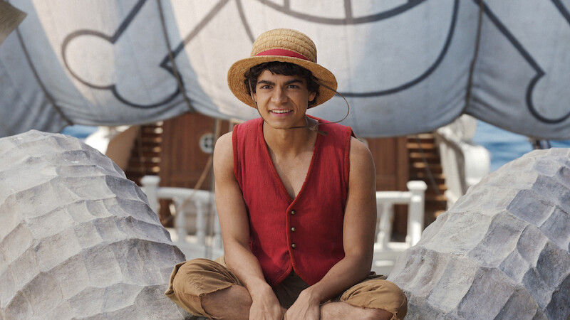 Luffy, a young man in a straw hat and red vest sits cross-legged on a ship's deck, with large decorative sails and nautical details in the background, suggesting a lively maritime adventure setting.