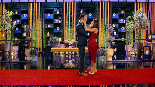 A couple stands on a red carpet, embracing in a decorated, modern indoor setting with potted plants, candles, and geometric décor on shelves in the background. Elegant, romantic atmosphere with formal attire.