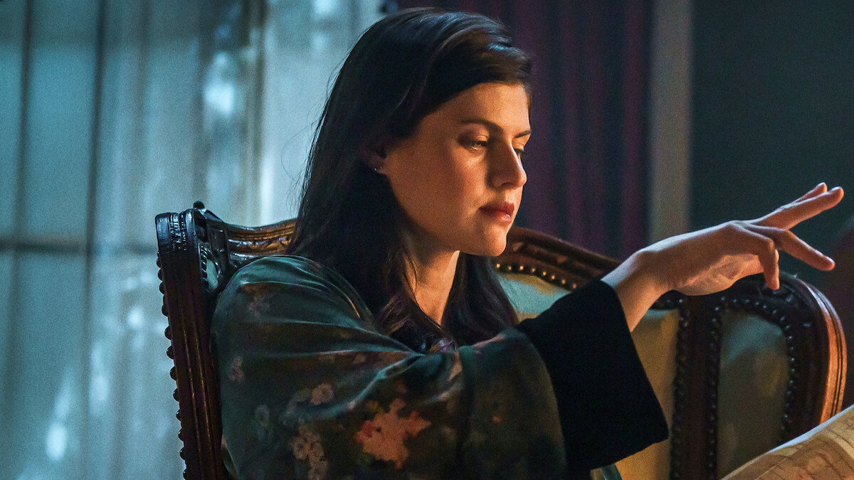 Woman sitting on ornate chair in dimly lit room, wearing a patterned robe, focused with her hand raised as if performing a gesture or casting a spell, with soft light coming through a window in the background.