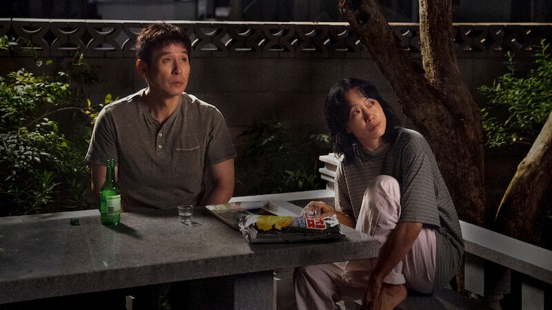 Two people sit at a stone table outdoors at night, surrounded by trees and plants, with snacks, a bottle, and a glass on the table, looking upward as if observing something in the sky.