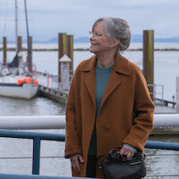 Sally Field as Tova in a brown coat standing by a railing at a marina, with sailboats docked and calm water in the background under a cloudy sky.
