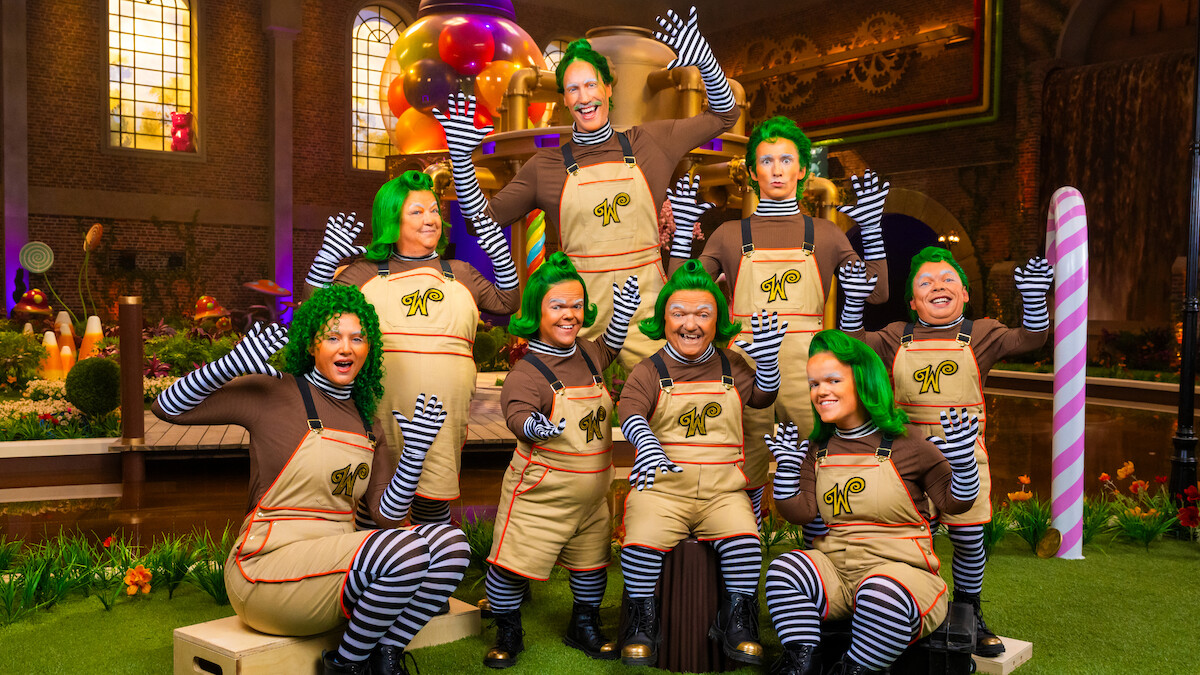 Grupo de pessoas fantasiadas de umpa-lumpas, com perucas verdes, camisas marrons, leggings listradas e macacões, no cenário de uma fábrica de doces colorida e extravagante, com guloseimas gigantes e iluminação vibrante.