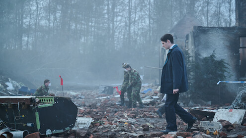 A man in a suit walks through a debris-strewn, foggy ruin as two people in camouflage uniforms investigate the wreckage; trees and a partially destroyed building are in the background.