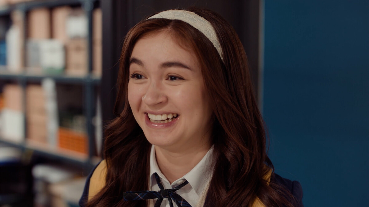 Smiling young woman with a headband and school uniform in an indoor setting, standing near blue wall and bookshelves.