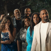A group of six adults dressed in formal and semi-formal attire stand close together outdoors at night, looking intensely ahead with concerned or serious expressions, trees and dark background behind them.