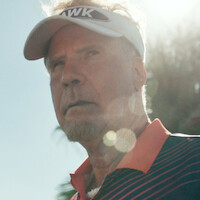 A man wearing a sports visor and striped shirt stands outdoors in the sunlight, with palm trees and blue sky in the background, giving a serious expression.