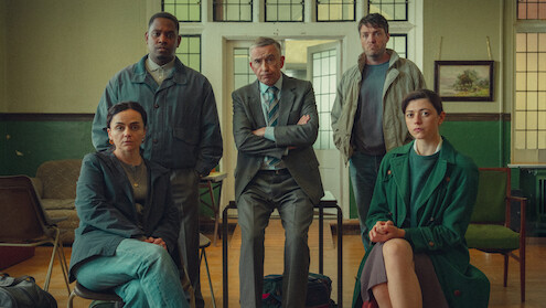 Five adults, three seated and two standing, pose in a serious group setting inside a classroom or office with green walls, wooden floor, chairs, and a painting on the wall, suggesting a meeting or discussion environment.