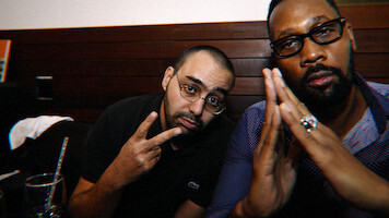 Two men sitting indoors at a table, one making a peace sign and the other with hands pressed together, both wearing glasses in a dimly-lit setting with wooden paneling in the background.