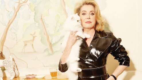 Woman in black leather outfit holding fluffy white cat, standing indoors by a painted wall with a deer mural, a table with tea, snacks, and decorative objects in a softly lit, stylish setting.