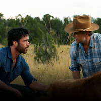 Two men kneel in a grassy field framed by trees. Yancy Grey (Jack Schumacher) wears a blue denim shirt, while Staten Kirkland (Josh Duhamel) sports a denim shirt and a cowboy hat.