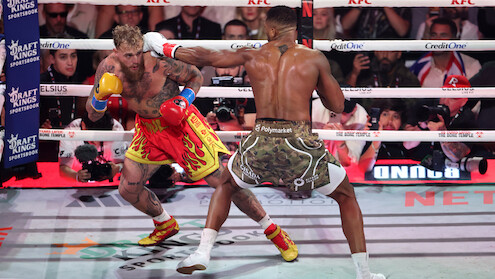 Jake Paul and Anthony Joshua fight in a brightly lit boxing ring, surrounded by ropes and an audience, with camera crews and sponsors visible in the background.