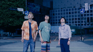 Three young men stand on a city crosswalk at night, looking up with curious or concerned expressions. The urban street is quiet and dimly lit, surrounded by buildings and street signs.