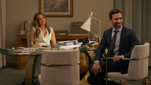 A woman and a man in business attire sit and smile in a modern office with a glass desk, papers, books, and a desk lamp, with neutral-colored walls and a framed picture in the background.