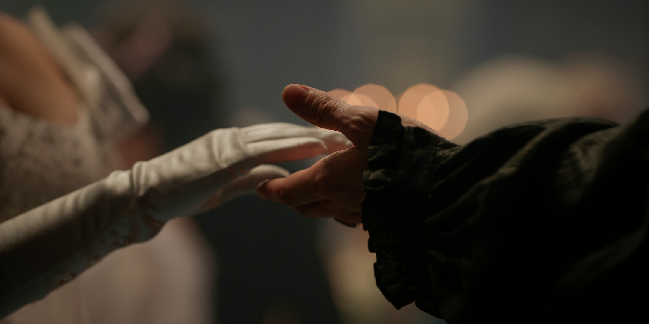 Two people reach toward each other, one wearing a white glove and elegant attire, the other in dark clothing, set in a softly lit, formal ballroom with blurred figures and warm lights in the background.