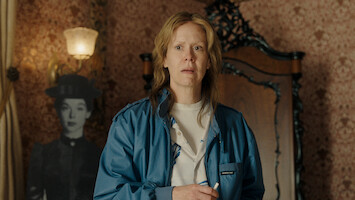 A concerned person in a blue jacket stands in a vintage room with patterned wallpaper, ornate wooden furniture, and a black-and-white cutout of a woman in period clothing in the background.