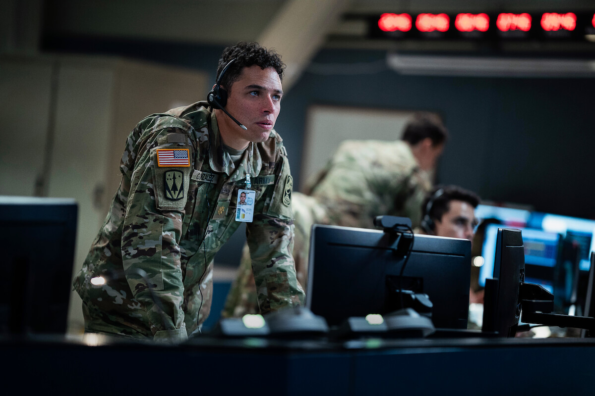 US military officer in camouflage with headset leans over monitor in command center, focused on screens; other soldiers work in background, intense and serious atmosphere.