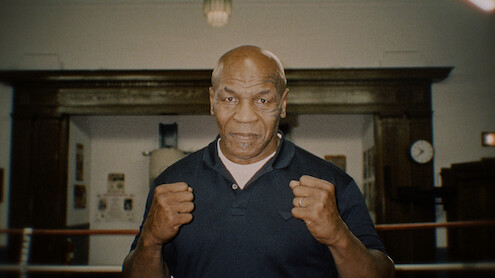 Man standing in a boxing gym with fists raised, wearing a dark shirt, wooden wall and clock in background, training environment with boxing ring ropes visible.