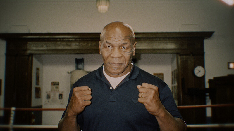 Man standing in a boxing gym with fists raised, wearing a dark shirt, wooden wall and clock in background, training environment with boxing ring ropes visible.