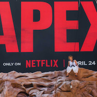 A person sits on artificial rocks in front of a large billboard for a Netflix show or movie, featuring bold red letters and a release date of April 24, creating a dramatic promotional scene.