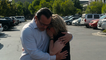 Two people embrace emotionally in a parking lot surrounded by cars and trees, suggesting comfort or consolation in a public, outdoor setting on a sunny day.