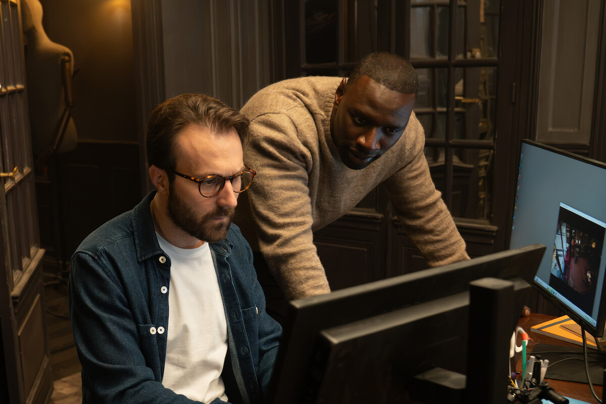 Um homem negro vestido com blusa bege está em pé, inclinado sobre uma mesa, ao lado de outro homem branco de camisa jeans e camiseta branca, barba e óculos, que está sentado, e ambos olham para a tela de um monitor.
