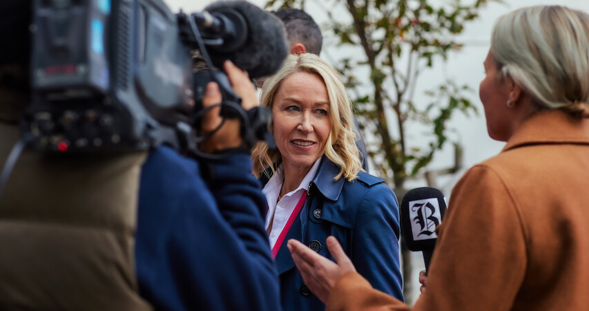 Trine Wiggen in a production still from the series 'Billionaire Island' faces the camera as reporters with a camera and microphone talk to her outside.