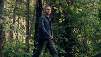 A man with gray hair and a beard walks through a dense, green forest wearing a dark jacket and jeans. The environment is lush with foliage, trees, and natural light filtering through the leaves.
