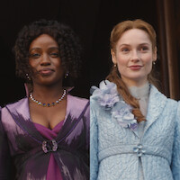 Two women in elegant period costumes, one in purple with a jeweled necklace and one in light blue with floral details, standing side by side against a dark background.
