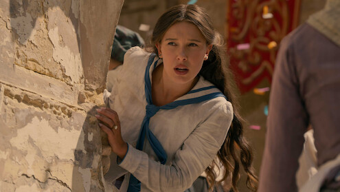 Young woman with long hair in historic clothing leans anxiously around a stone wall in an outdoor setting, with colorful confetti in the air and other people nearby, suggesting a dramatic or festive event.
