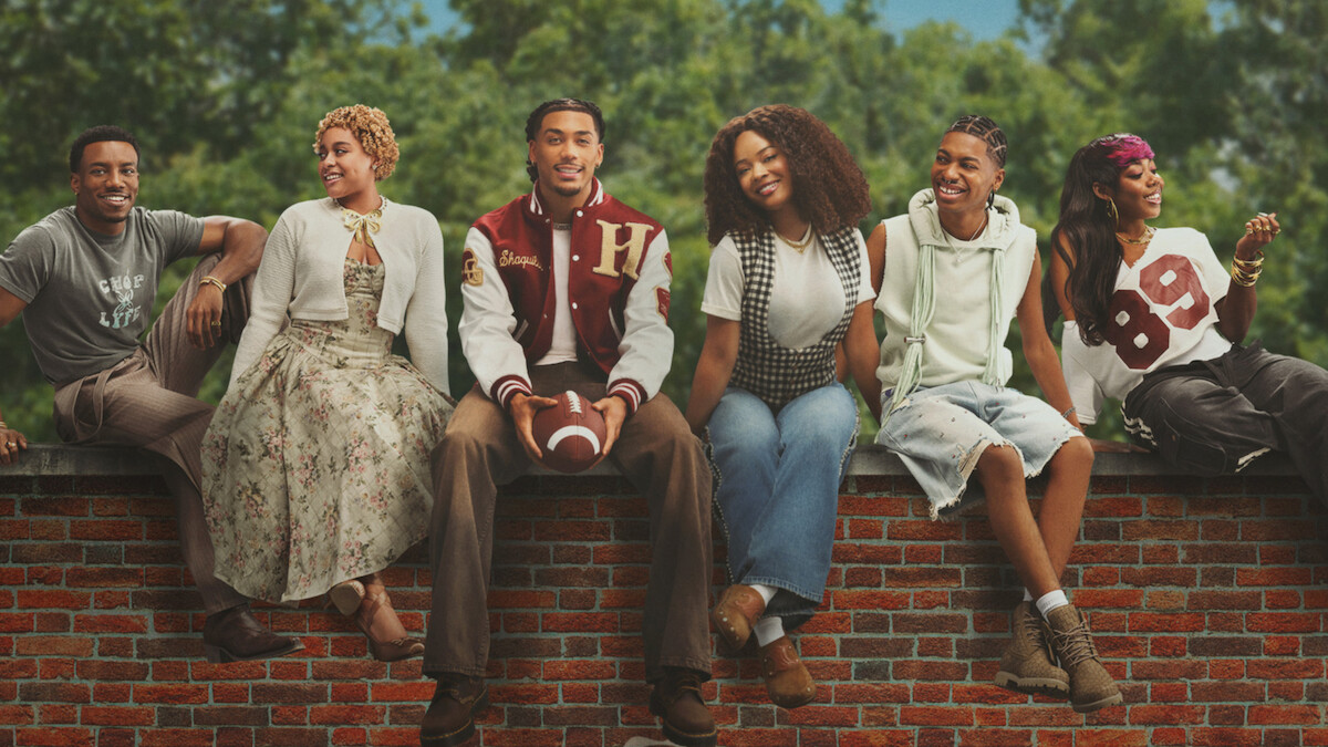 Six young adults sit on a brick wall outdoors, smiling and relaxed. Trees and blue sky in background. One holds a football; they wear casual, varied outfits, indicating a friendly, laid-back group setting.