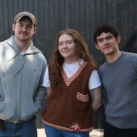Three young adults standing outside in casual clothing, smiling in front of a dark vertical wooden fence on a bright day.