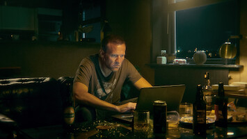 A man sits in a dimly lit, cluttered room at night, focusing intently on his laptop. The environment feels tense and isolated, with bottles and plates scattered around a table, suggesting loneliness or stress.