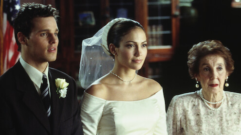 A bride in a white dress and veil stands between a man in a suit with a boutonniere and an elderly woman in pearls, indoors with bookshelves and an American flag in the background.