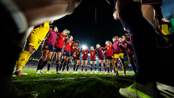 A women's soccer team huddles together on a grass field at night under stadium lights, wearing matching uniforms, preparing for a match with an atmosphere of unity and focus.