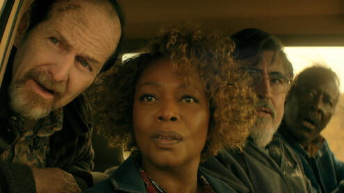 Four adults sit closely together inside a car, looking intently out the window. The lighting is warm, and the expressions on their faces suggest concern or curiosity. The car interior and a glimpse of the outdoors are visible.
