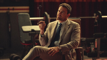 Man in a tan suit sitting in a chair indoors, holding a cigar and looking thoughtful. The background features gym or fitness equipment and dim, moody lighting.