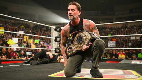 CM Punk kneels in a wrestling ring holding a championship belt, with a cheering crowd and colorful signs in the background during a live event.
