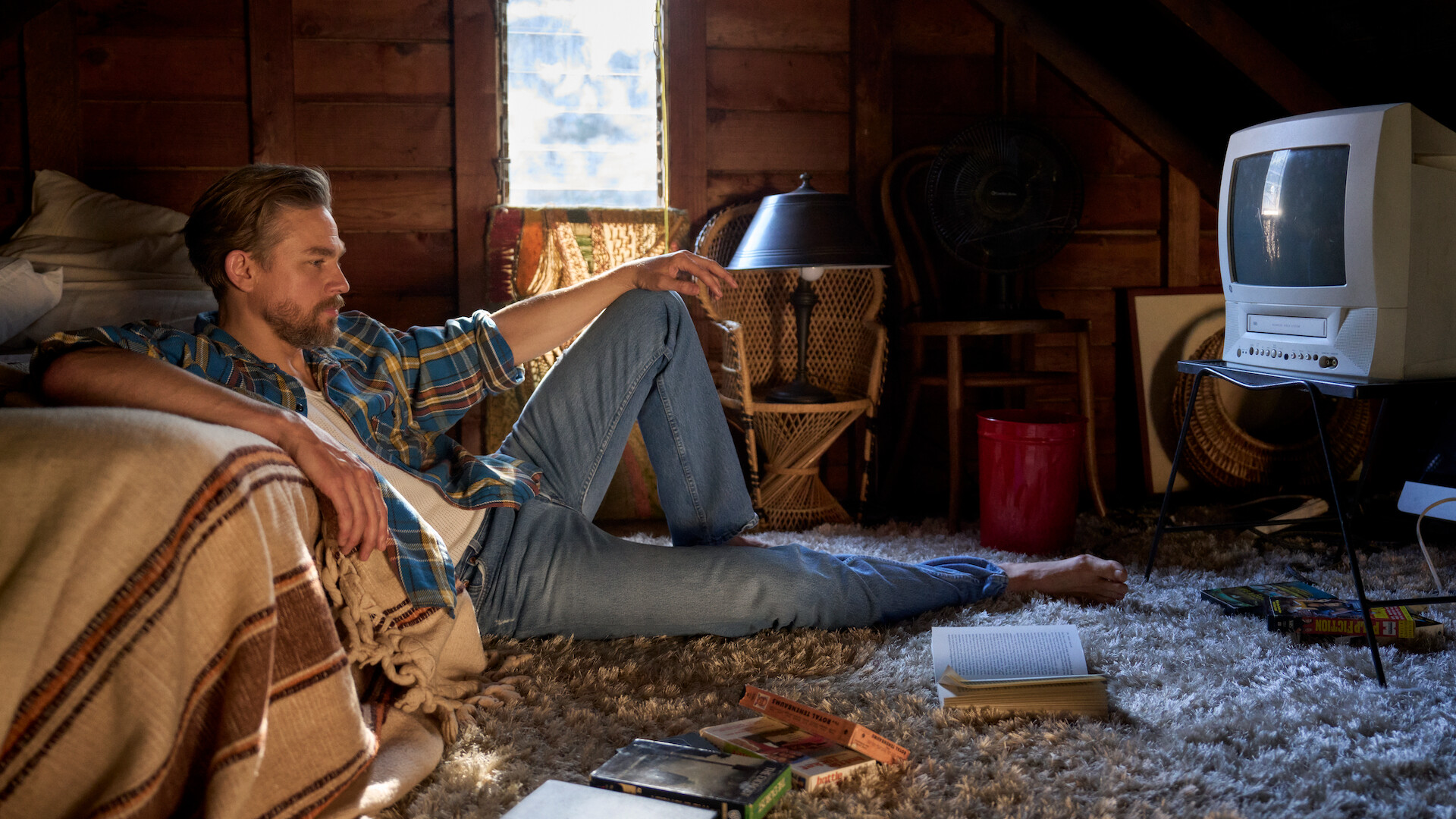 Charlie Hunnam sits in a cabin bedroom on the floor surrounded by VHSs and books, staring into the distance with a contemplative look on his face. 