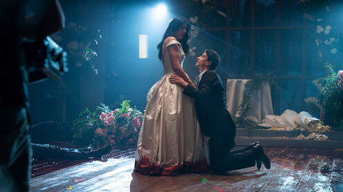 A man in a suit kneels before a woman in a white dress with red trim in a dimly lit, flower-filled room, dramatic lighting highlighting the emotional scene, with petals and decorations scattered on the wooden floor.