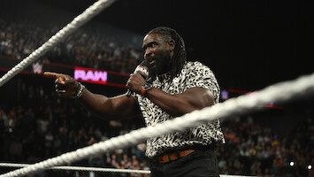 A man with a microphone stands in a wrestling ring, pointing as he speaks to a large crowd during a WWE Raw event in an arena.
