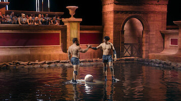 Two men in athletic gear stand in a shallow arena pool, shaking hands next to a ball, with spectators watching from seats above in a setting resembling ancient Roman architecture.