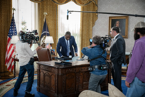 Actor in a suit stands at a presidential desk in an Oval Office set, surrounded by film crew, cameras, and lighting equipment, creating a busy, professional atmosphere for a political drama scene.