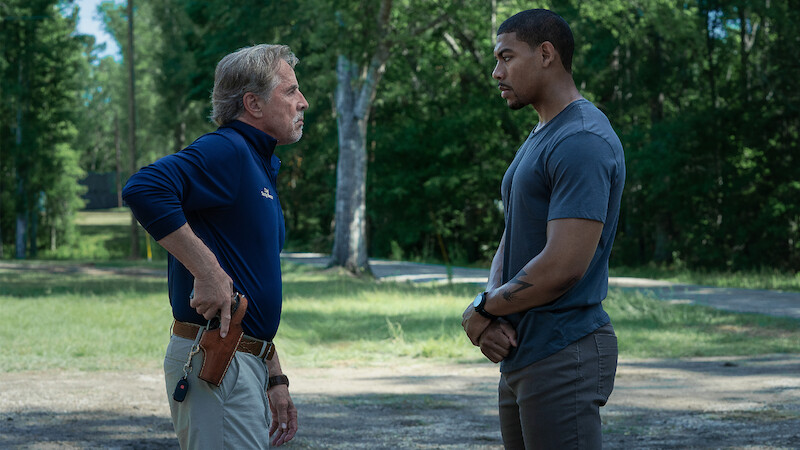 Two men stand outdoors facing each other in a grassy, wooded area. One man, older and wearing a blue shirt with khaki pants, has a hand on a holstered gun. The other, younger man, stands calmly with hands clasped.