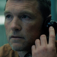 Close-up of a man with short hair and stubble holding a black telephone receiver to his ear, appearing thoughtful. He is indoors, possibly in a serious or intense setting with muted lighting and a blue background.